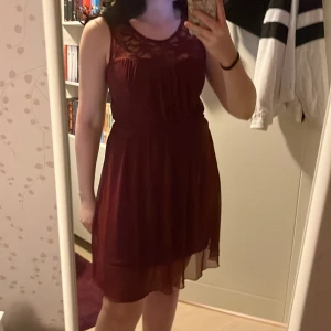 Vinröd ärmlös klänning med spets - Vinröd ärmlös klänning med vacker spets upptill och rund halsringning. Kjolen är lätt och faller snyggt, och ryggen har en liten öppning med knapp. 