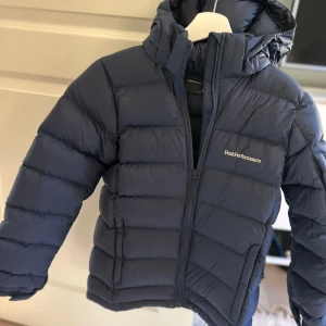 Mörkblå dunjacka Peak Performance Frost down stl 160 - Snygg mörkblå dunjacka från Peak Performance med huva och dragkedja. Jackan är quiltad och har logga på bröstet samt ärmen. Perfekt för kalla dagar och har en stilren, sportig look.Modellen heter Frost down jacket . Nypris 2500 kr. 