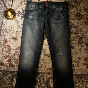 Slim Glenn jeans från Jack & Jones - Snygga mörkblå jeans från Jack & Jones, modell Slim Glenn. Jeansen har slim fit-passning, knäppning med flera knappar och slitna detaljer på låren och knäet. Klassisk femficksdesign och cool faded effekt. Perfekta för dig som gillar en modern och avslappnad stil.