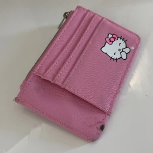 Hello Kitty plånbok från Bershka - Söt rosa Hello Kitty plånbok från Bershka med flera kortfack och dragkedja. Plånboken har tryck med Hello Kitty-figurer, små stjärnor och hjärtan. Tillverkad i syntetmaterial och perfekt för dig som gillar gulliga accessoarer.
