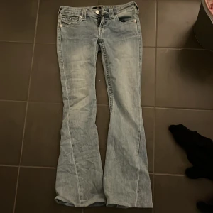 True Religion Joey ljusblå flare jeans - Säljer ett par ikoniska True Religion Joey jeans i ljusblå tvätt med låg midja och flare-ben. Klassiska fickor med knappar bak och snygga detaljer som synliga sömmar och logopatch. Perfekt för dig som gillar retrovibe och statement-jeans. Säljer mina helt nya jeans då dem är lite för stora i midjan 