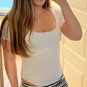 Vit tight kortärmad body - Säljer en vit, tight och kortärmad body med djup fyrkantig ringning. Toppen är figurnära och har en clean, minimalistisk look som är enkel att matcha. Materialet känns mjukt och stretchigt, perfekt för dig som gillar en slim fit och enkel stil.