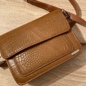 Brun axelväska i croco-mönster från Cocopat - Snygg brun axelväska från Cocopat med croco-mönstrad yta och justerbar rem. Väskan har flera fack inuti och stängs med lock. Perfekt storlek för det viktigaste och ger en trendig touch till din outfit.