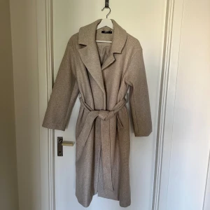 Beige kappa med bälte från Gina tricot - Stilren beige kappa från Gina Tricot med klassisk krage och knytskärp i midjan. Lång modell med raka ärmar och mjukt ullmaterial som ger en lyxig känsla. Passar både dig med storlek xs/s/m. Perfekt för dig som vill ha en tidlös och snygg look under kyliga dagar.