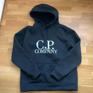 Svart hoodie från C.P. Company - Svart hoodie från C.P. Company Stilren svart hoodie med stor vit logga på bröstet. Mjukt och bekvämt material med klassisk passform. Bra till vintern.