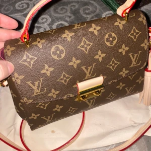 L väska  - Snygg axelväska i klassiskt monogrammönster. Väskan är brun med beige och guldiga detaljer, har ett handtag och avtagbar axelrem med röda kanter. Insidan är fodrad i vinrött tyg och väskan stängs med ett guldfärgat lås framtill. Öppen för prisförslag men ej skambud. Väskan är inte använt helt ny säljes pga fel köp av model. 