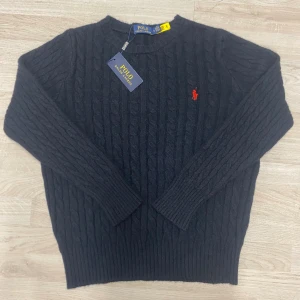 Svart kabelstickad tröja Polo Ralph Lauren - Svart kabelstickad tröja från Polo Ralph Lauren med klassisk röd broderad logga på bröstet. Tröjan har rund hals, ribbade muddar och är långärmad. Materialet är mjuk ull som ger en skön känsla och snygg passform.
