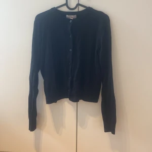 Svart kofta från H&M, storlek M/170 - En stilren svart kofta från H&M i storlek M. Koftan har rund hals, långa ärmar och knäppning med svarta knappar framtill. Materialet känns mjukt och lätt, perfekt att slänga på sig när det är lite kyligt ute.