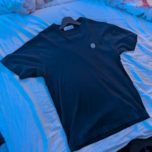 Svart t-shirt från Stone Island - Svart t-shirt från Stone Island med klassisk rund hals och korta ärmar. På bröstet sitter den ikoniska Stone Island-loggan i svart och vitt. T-shirten är gjord i mjuk bomull och har en clean, stilren look som passar till jeans eller shorts. 
