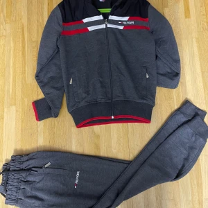 Grå zip hoodie från Tommy Hilfiger - Snygg mörkgrå hoodie med dragkedja från Tommy Hilfiger, med röda och vita detaljer på bröstet och logga på sidan. Långärmad med ribbade muddar och röd kant vid ärmslut och nederkant. Perfekt för chill eller träning.