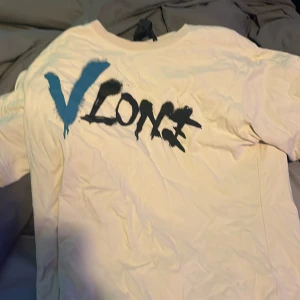Vit VLONE t-shirt med blått tryck - Säljer en vit t-shirt från VLONE i storlek L med stort blått V-tryck på ryggen och graffiti-inspirerad VLONE-logga i blått och svart på bröstet. T-shirten har korta ärmar och är gjord i mjuk bomull. Perfekt för dig som gillar streetwear och vill sticka ut.
