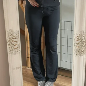 Svarta bootcut byxor med snörning - Säljer ett par svarta bootcut byxor från Nly trend  i storlek S. Byxorna har hög midja, snörning framtill och är gjorda i ett glansigt, stretchigt material som sitter tajt upptill och släpper ut vid benen. Perfekta för en trendig och bekväm look.