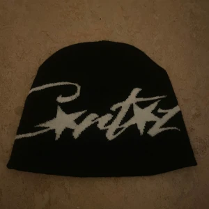 Svart mössa från Carltz med vit logga - Svart mössa med vit, stor Carltz-logga i graffiti-stil och stjärndetaljer. Mjuk och stretchig, perfekt för kalla dagar. Snygg streetwear-vibe som lyfter din outfit. Materialet är stickat och färgerna är svart och vit.