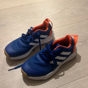 Blå adidas sneakers (t.ex. handbollsskor) med orange detaljer - Snygga blå sneakers (tex handbollsskor)  från adidas med vita ränder på sidorna och orange insida samt hälflik. Ovandelen är i ventilerande syntetmaterial och sulan är platt med gummiyta för bra grepp. Perfekta för dig som gillar sportig stil och färgkombinationer.