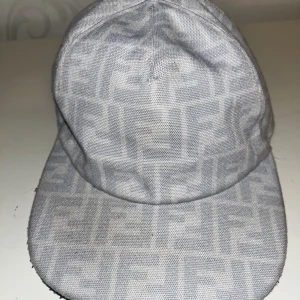 Grå Fendi keps med logomönster - Säljer en snygg keps från Fendi i ljusgrått med det klassiska FF-logomönstret över hela kepsen. Kepsen är tillverkad i bomull och har justerbar rem baktill. Perfekt accessoar för att lyfta din outfit med en exklusiv touch.