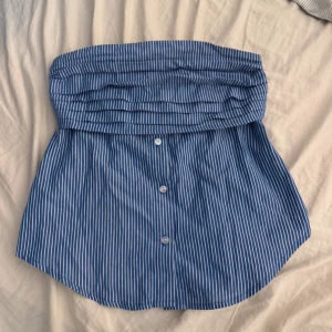 Blåvit randig bandeau tubtopp - Säljer en blå och vit randig bandeau tubtopp från FB Sister. Toppen är axelbandslös med knappar framtill och dragkedja i sidan för enkel påklädning. Tillverkad i mjuk bomull, perfekt för varma dagar. Snygg och chill stil som passar till jeans eller shorts.