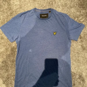 Blå t-shirt från Lyle & Scott - Snygg blå t-shirt från Lyle & Scott med klassisk rund hals och korta ärmar. På bröstet sitter den ikoniska gula fågeln som broderad detalj. Materialet är en skön mix av bomull och polyester som känns lätt och bekvämt mot huden.