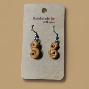 Lussebulle - Unika handgjorda örhängen som ser ut som små lussebullar, med snirklig form och detaljerade russin. Färgen är gyllene gul med bruna inslag, och krokarna är i silverfärgad metall. Perfekt för dig som vill sticka ut med något kreativt och roligt.