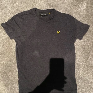 Mörkgrå t-shirt från Lyle & Scott - Snygg mörkgrå t-shirt från Lyle & Scott med klassisk rund hals och korta ärmar. På bröstet sitter den ikoniska gula fågel-loggan som ger en clean och stilren vibe. Tillverkad i 100% bomull för skön komfort hela dagen.