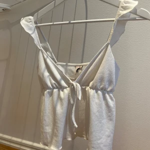 Vit linnetopp med volang och knyt - Supersöt vit linnetopp med volangdetaljer på axelbanden och knyt framtill. Toppen har en v-ringad design och är croppad med lätt rynk vid midjan. Perfekt för varma dagar och har en mjuk, lite texturerad bomullskänsla.