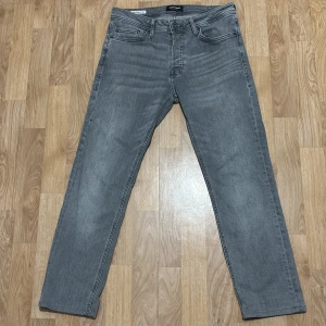 Grå slim straight jeans Jack & Jones - Slim straight jeans från Jack & Jones i snygg grå tvätt. Klassisk femficksmodell med knapp och dragkedja fram. Modellen heter Tim och har en rak passform som sitter schysst på benen. Perfekt för dig som gillar stilrena och enkla jeans.