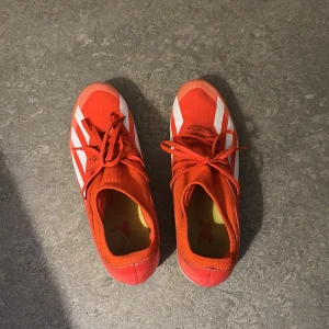 Adidas Crazyfast röda fotbollsskor storlek 45 1/3 - Säljer ett par Adidas Crazyfast fotbollsskor i röd färg med vita ränder och gula dobbar. Skorna har snörning och en strömlinjeformad design med rund tå. Ovandelen är i syntetmaterial och insidan är gul. Perfekt för dig som vill ha snabba och snygga skor på planen.