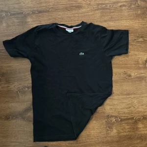 Svart t-shirt från Lacoste - Svart t-shirt från Lacoste med klassisk rund hals och det ikoniska gröna krokodilmärket broderat på bröstet. T-shirten är i mjuk bomull och har en enkel, stilren design med korta ärmar. Perfekt för en clean och avslappnad look.