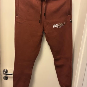 Bruna Hoodrich mjukisbyxor med snörning - Snygga bruna mjukisbyxor från Hoodrich med vit logga på benet och broderad text. Byxorna har snörning i midjan, smal passform och praktiska fickor med dragkedja. Perfekta för chill eller streetwear-stil.
