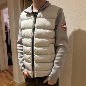 Ljus pufferjacka från Canada Goose M - Snygg ljus pufferjacka från Canada Goose med quiltad front och släta ärmar. Jackan har dragkedja, två fickor med knappstängning och klassisk logga på ärmen. Materialet är bomull och polyester, perfekt för kyliga dagar. Köptes för ca 3800kr på plick. Passar även S sitter fint se sista bilden killen på bild är S och ca 1.78