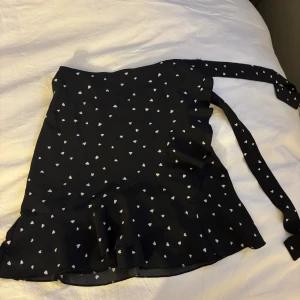 Svart omlottkjol med vita hjärtan - Supersöt svart kort omlottkjol med vita hjärtan och volangkant. Kjolen knyts i sidan och har ett lekfullt mönster som verkligen sticker ut. Perfekt för dig som gillar gulliga detaljer och vill ha en kjol som känns både trendig och unik.