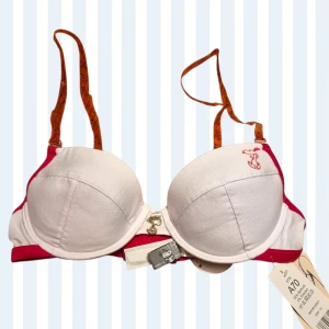 Ny vit och röd Snobben-bh 70A - Supersöt linnetopp från Snoopy med vita kupor och röd kant. Axelbanden är transparenta orange med Snoopy-tryck. Materialet är 95% bomull och 5% elastan för skön stretch. Perfekt för dig som gillar roliga detaljer och mjuka färger.