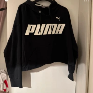 Svart hoodie från Puma med logga - Svart hoodie från Puma med stor vit logga framtill. Tröjan har huva med snörning och en klassisk känguruficka. Croppad passform och långa ärmar ger en chill och sportig vibe. Perfekt för dig som gillar streetwear och enkel stil.