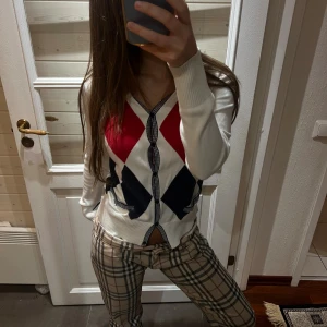 Argyle kofta från Tommy Hilfiger - Snygg kofta från Tommy Hilfiger med argylemönster i rött, vitt och marinblått. V-ringad med knappar framtill och två små fickor. Långärmad och tillverkad i mjuk bomull, perfekt för lager-på-lager-stil.