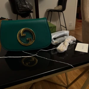 Grön axelväska från Gucci i skinn - Snygg grön axelväska från Gucci i slätt skinn med stort guldfärgat GG-spänne framtill. Väskan har en stilren, rundad form och levereras med originalkartong och axelrem. Perfekt för dig som vill sticka ut med en lyxig accessoar.