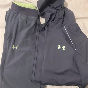  tracksuit från Under Armour - Säljer en grå hoodie från Under Armour med limegröna loggor och detaljer. Tröjan har huva, dragkedja framtill och är tillverkad i ett lätt och sportigt syntetmaterial. Perfekt för träning eller chill. Snyggt tryck på ryggen och ärmarna.