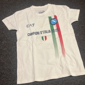 Vit Napoli EA7 t-shirt Campioni 22/23 - Vit t-shirt från EA7 x Napoli med tryck 'Campioni d'Italia 22/23' och italienska flaggfärger framtill. Stor guldig 3:a och 'Campioni' på ryggen. Crewneck, kortärmad och i 100% bomull. Perfekt för dig som älskar fotboll och vill hylla Napolis mästartitel.