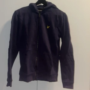 Mörkblå hoodie från Lyle & Scott - Mörkblå hoodie från Lyle & Scott med dragkedja framtill och klassisk gul logga på bröstet. Tröjan har huva, fickor och långa ärmar. Perfekt för en chill och stilren look.