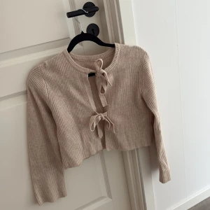 Beige ribbad kofta med knyt - Superfin beige ribbad kofta med croppad passform och två knyt framtill. Långärmad och i mjukt stickat material, perfekt att slänga över ett linne eller t-shirt. Enkel och stilren med en trendig twist.