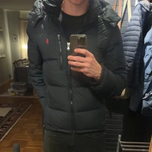 Svart dunjacka från Polo Ralph Lauren - Säljer en svart dunjacka från Polo Ralph Lauren med röd logga på bröstet. Jackan är knappt använd och har knappt något tecken på användning. Perfekt för kalla dagar och har en snygg, modern look med lång ärm och normal passform. Pris kan diskuteras vid snabbaffär. Jackan är köpt för 4000 kr så mitt pris är perfekt för dig som inte vill lägga ut en stor summa pengar! Vid funderingar tveka inte att kontakta mig🤩🙌