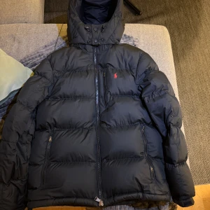 Svart pufferjacka från Polo Ralph Lauren - Svart pufferjacka från Polo Ralph Lauren med huva och dragkedja framtill.Köpt från Boozt, använd 1 säsong. Nypris runt 5000kr. Inga defekter alls, vädligt fint skick! 
