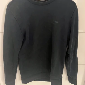 Svart sweatshirt från BOSS - Svart sweatshirt från BOSS med diskret logga på bröstet. Tröjan har rund halsringning och långa ärmar med ribbade muddar. Materialet är mjuk bomull som ger en skön och avslappnad känsla. Perfekt för dig som gillar stilrena och enkla plagg.