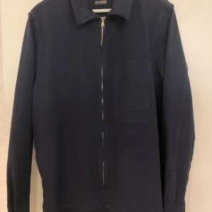 Mörkblå jacka från C.O.F. Studio - Stilren mörkblå jacka från C.O.F. Studio med klassisk krage, bröstficka och hel dragkedja framtill. Långärmad modell, perfekt att bära som lager över t-shirt eller hoodie.