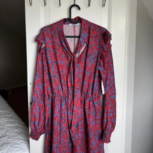 Röd blommig klänning med volang - Röd långärmad klänning med blått blommönster. Klänningen har volangdetaljer på axlarna, knytband vid halsen och smockade ärmslut. Ny med prislapp 