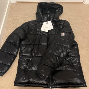 Svart Moncler pufferjacka med huva - Svart pufferjacka från Moncler med glansig finish, huva och dragkedja framtill. Jackan har Moncler-logga på ärmen, två sidofickor med dragkedja och coolt serietryck på insidan. Perfekt för dig som gillar streetstyle och vill hålla dig varm.