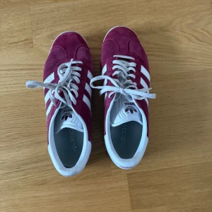 Adidas Gazelle vinröda sneakers - Säljer ett par Adidas Gazelle sneakers i vinrött med klassiska vita ränder och vit häl. Lätt använda men väl skyddade och fortfarande väldigt bra skick!