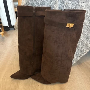 Bruna mockastövlar med spetsig tå - Säljer ett par höga bruna stövlar i mocka med spetsig tå och låg klack. Stövlarna har dragkedja på insidan och en snygg guldfärgad detalj upptill. Perfekta till höst- och vinterlooken och passar till både kjol och byxor.