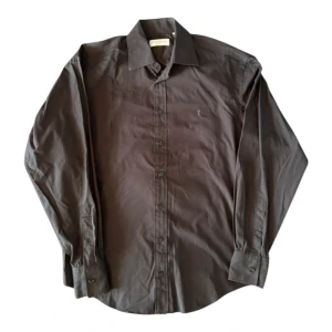 Svart skjorta från Yves Saint Laurent - Snygg svart skjorta från Yves Saint Laurent med klassisk krage och knappar hela vägen. Diskret YSL-broderi på bröstet ger en exklusiv touch. Tillverkad i mjuk bomull som känns skön mot huden. Perfekt för dig som gillar stilrena designer och vill ha något från ett ikoniskt märke. Storlek 39, borde passa M-L