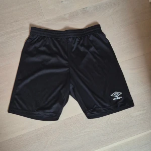 Svarta träningsshorts från Umbro L - Svarta träningsshorts från Umbro i lätt och glansig polyester. Klassisk sportig look med elastisk midja och logga på ena benet. Perfekta för fotboll eller gymmet, riktigt sköna och enkla att matcha med din favorit-t-shirt. Nästan oanvända.
