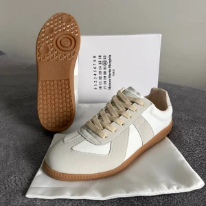 Maison Margiela skor  - Maison Margiela  sneakers i vit och beige med klassisk gummisula i brunt. Skorna har snygga paneler i skinn och mocka, rund tå och beige snörning. Ikonisk logga på plösen och stilren design som ger en clean vibe.