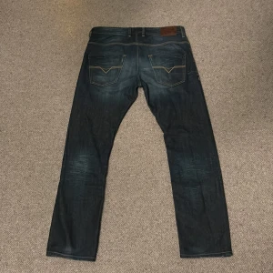 Diesel DNA mörkblå jeans straight fit - Säljer ett par snygga mörkblå jeans från Diesel med coola slitningar och kontrastsömmar. Modellen har raka ben, klassiska fem fickor och detaljer som orange DNA-tryck i midjan samt Diesel Industry-märke vid fickan. Perfekt för dig som gillar streetstyle. Storlek W34 L32. Pris kan diskuteras.